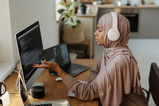 Young Muslim female programmer pointing at data on computer screen while sitting by workplace and communicating with colleague in video chat - Powered by Adobe