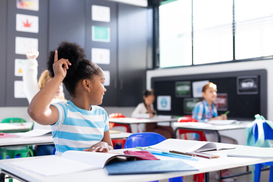 African american schoolgirl sitting at desk raising hand in elementary school classroom, copy space