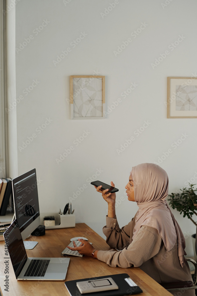 Poster young businesswoman in hijab recording voicemail on smartphone while sitting by workplace in front o