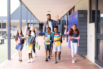 Diverse, happy male teacher and elementary schoolchildren waving in school corridor, copy space - Powered by Adobe