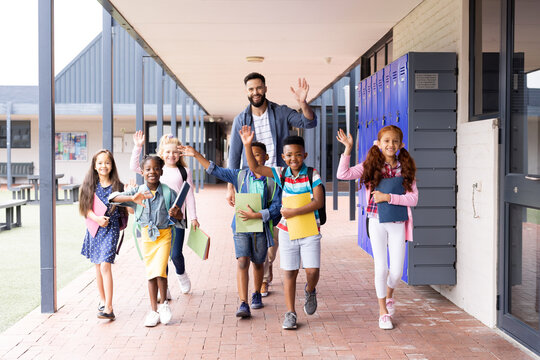 Diverse, Happy Male Teacher And Elementary Schoolchildren Waving In School Corridor, Copy Space