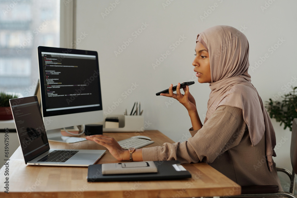 Poster side view of confident muslim female programmer talking by speakerphone during work while holding mo