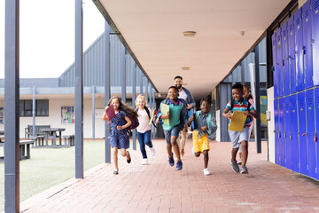 Diverse male teacher with happy children running in elementary school corridor, copy space