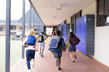Rear view of diverse children with schoolbags running in elementary school corridor, copy space
