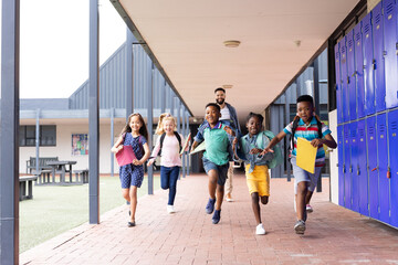 Happy, diverse male teacher with children running in elementary school corridor, copy space