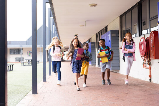 Diverse male teacher with happy children running in elementary school corridor, copy space