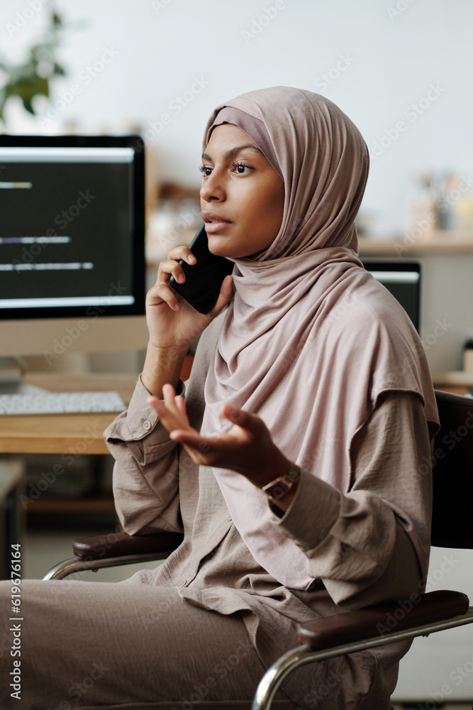 Poster young confident female it engineer or programmer in hijab talking on smartphone while sitting by wor