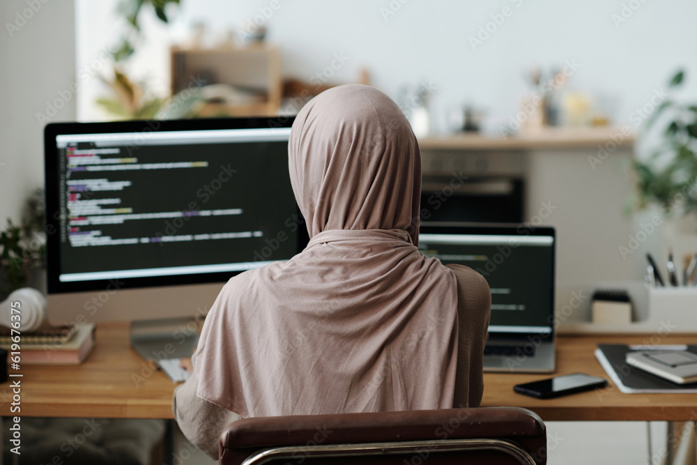 Poster Rear view of Muslim female programmer in hijab sitting in front of computer screen with coded data and using new decoding software