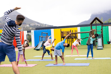 Diverse male teacher and elementary schoolchildren practicing yoga in schoolyard