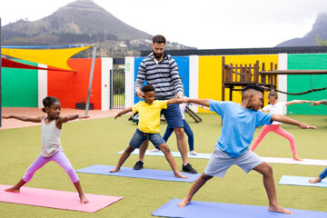 Diverse male teacher and elementary schoolchildren learning yoga in schoolyard