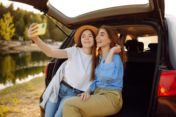 Smiling young womans have fun in the back seat of a car while traveling. Girlfriends take selfies,...