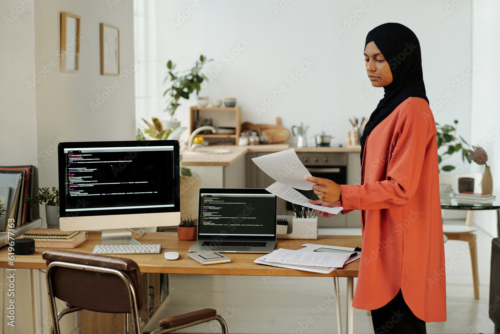 Aufkleber young muslim female programmer or accountant looking through papers while standing by workplace with
