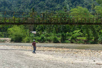 Man standing near the river with old hanging bridge, in Lokop Serbajadi, east Aceh, Indonesia