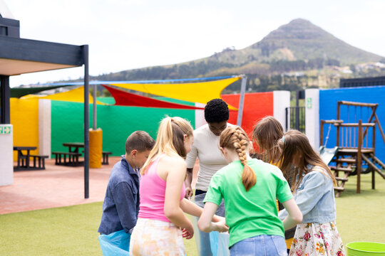 Diverse female teacher and pupils collecting rubbish for recycling in schoolyard, with copy space