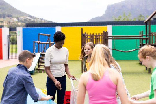 Diverse female teacher and pupils collecting rubbish for recycling in schoolyard, with copy space