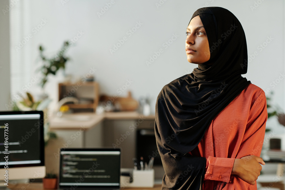 Poster young serious businesswoman in black hijab looking aside while standing in front of camera against w