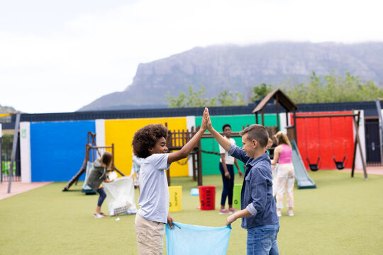 Two diverse schoolboys collecting rubbish for recycling high fiving in schoolyard, with copy space