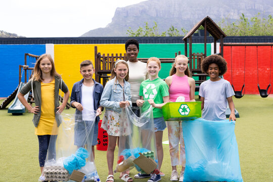 Portrait of happy diverse female teacher and pupils holding recycling collected in schoolyard