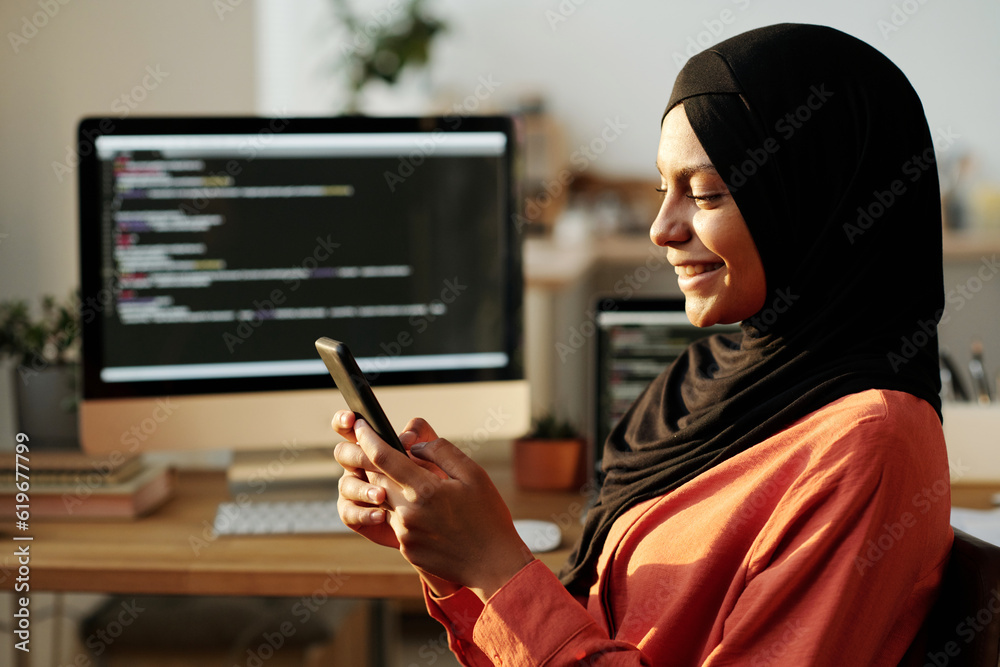 Poster side view of young cheerful muslim businesswoman watching video in smartphone while sitting in front
