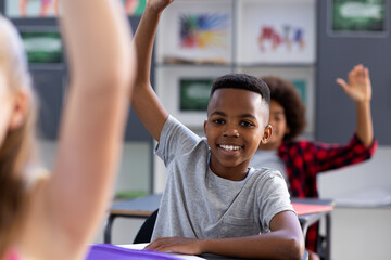 Portrait of smiling african american schoolboy sitting at desk raising hand in class