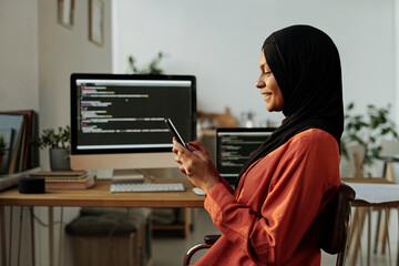 Young smiling Muslim woman looking at smartphone screen while texting, watching online video or communicating in video chat against workplace