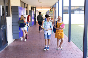 Smiling caucasian of boy and girl holding books, walking in school corridor talking with copy space
