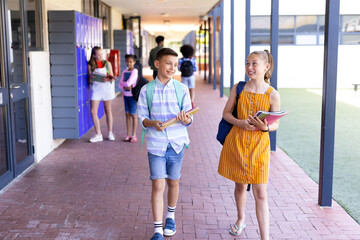 Smiling caucasian of boy and girl holding books, walking in school corridor talking copy space
