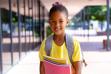 Happy biracial schoolgirl with school bag holding books, smiling outside school, with copy space