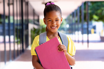 Happy biracial schoolgirl with school bag holding books, smiling outside school, with copy space