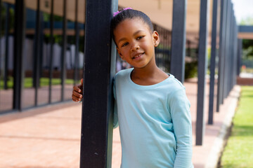 Portrait of happy biracial schoolgirl smiling, leaning on post in schoolyard copy space