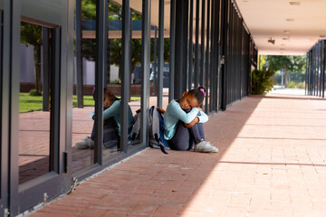 Sad, biracial schoolgirl with school bag sitting on the ground outside school, with copy space