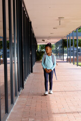 Vertical image of biracial schoolgirl with school bag walking outside school, with copy space