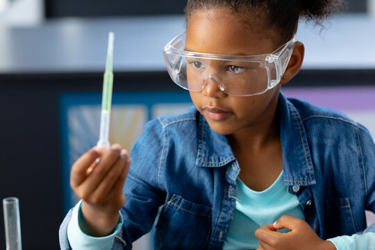 Happy biracial schoolgirl in safety glasses observing pipette in science class with copy space - Powered by Adobe