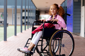 Portrait of happy biracial schoolgirl in wheelchair, smiling in school corridor