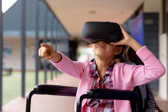 Biracial schoolgirl in wheelchair, using vr headset and pointing in school corridor