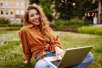 Beautiful female freelancer sits on a green meadow with a laptop. Online education. Happy woman running outdoors enjoying the sun. Freelancing concept.