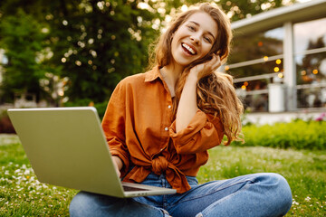 Beautiful female freelancer sits on a green meadow with a laptop. Online education. Happy woman running outdoors enjoying the sun. Freelancing concept.