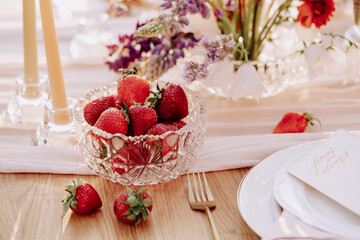 Strawberries in Bowl Standing on Dining Table Close-up Photography. Vitamin Fresh and Ripe Berries in Glass Dish and Empty Plate, Fork Utensil and Natural Flowers Bouquet on Wooden Desk