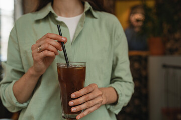 Close-up of woman in green linen shirt drinking summer refreshing beverage. A woman mixing espresso tonic with a straw.
