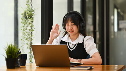 Smiling asian female student wearing headphones learning online, watching webinar class on laptop