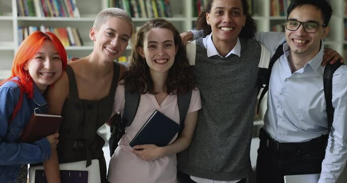 Happy young diverse student girls and guys looking at camera, smiling, laughing. Cheerful multiethnic teenage classmate posing in library, standing in row, hugging, speaking