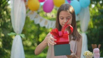 Portrait of surprised birthday girl opening gift making excited facial expression. Happy Caucasian child celebrating holiday on sunny summer day in park. Slow motion