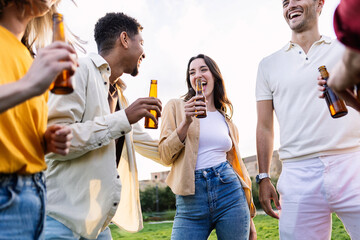Group of young friends celebrating together cheering with beer bottles outside. Multiracial happy people laughing enjoying summer party outdoors. Community, youth and friendship concept.
