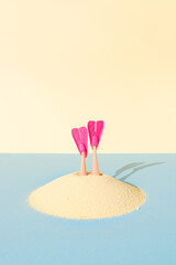 A pair of diving fins pokes out of the sand on blue and yellow pastel background. Creative idea of summer swimming recreation, or undersea vacation concept.