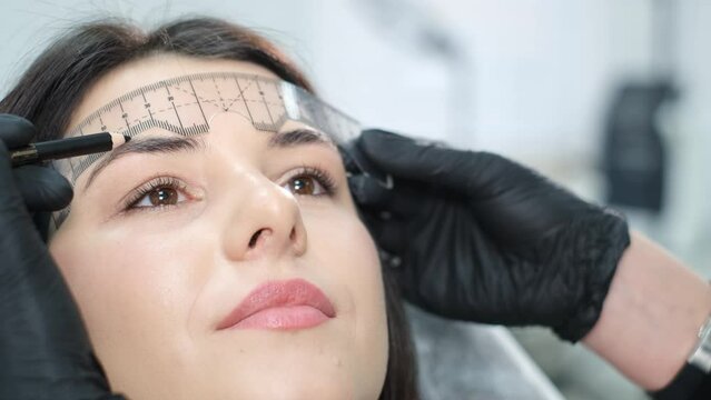 A close-up beautician corrects the shape of the eyebrows with a special ruler. Beautiful girl 