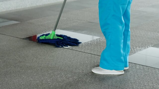 Close-up legs maid who cleans in one place, was using mop dirty floor, is rubbing shiny glass area clean in order give beautiful shine wearing blue pants, which is uniform cleaning staff.