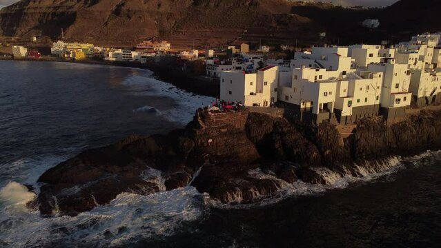 San Andres beach in Arucas: aerial view with movement in distance to the white buildings on the beach of San Andres and the waves hitting the coast. During sunset on the island of Gran Canaria.
