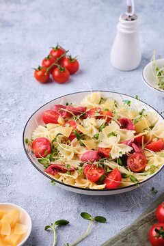 Pasta salad with farfalle pasta, cherry tomatoes, smoked duck meat and microgreens in a white plate on a light concrete background. Italian Cuisine.