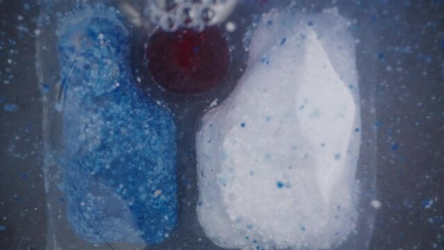The soluble packet for the dishwasher, with red and blue powder inside, dissolves under water, and the red gel slowly oozes out.