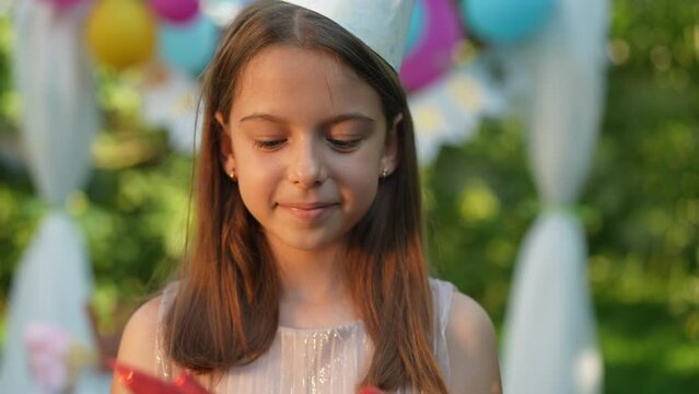 Satisfied pretty Caucasian girl looking at camera and admiring gift box in slow motion. Portrait of happy cute child posing outdoors with present as live camera zoom out